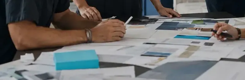 Hands reviewing printed layouts and color swatches laid out across a worktable